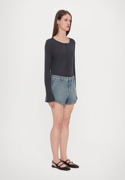 Top de manga larga gris oscuro con botones en el frente, combinado con pantalones cortos de mezclilla azul claro. La modelo lleva sandalias planas negras con tiras.