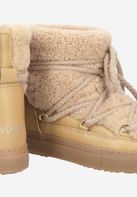 Bottes en cuir beige avec un col en fourrure tan duveteuse, dotées de lacets tressés et d'accents dorés, conçues pour la chaleur et le confort.