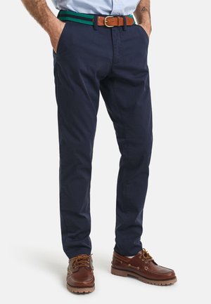 Marineblaue Baumwollhosen mit standardmäßiger Passform, ausgestattet mit einem grün-gestreiften gewebten Gürtel und braunen Lederakzenten. Braune Bootsschuhe runden den Look ab.