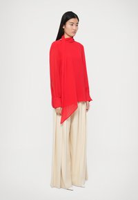 Blouse rouge à manches longues avec col montant et détail d'écharpe drapée, associée à un pantalon large beige, présentant une texture lisse et des poignets discrets.