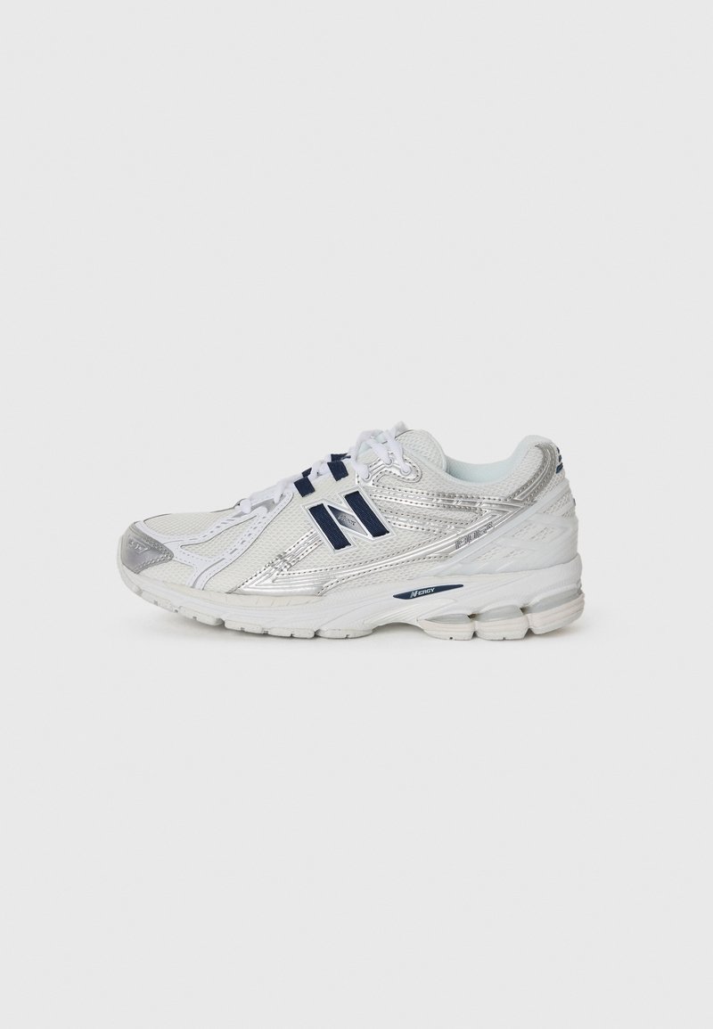 Chaussure de sport blanche et argentée avec logo New Balance bleu, fermeture à lacets et semelle amortissante sur un fond gris clair uni.