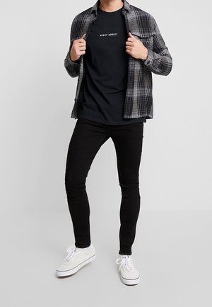 Homme portant un t-shirt noir "Night Addict", une chemise grise à carreaux déboutonnée, un jean noir slim et des baskets blanches sur un fond uni.