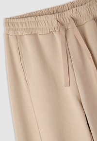 Pantaloni della tuta beige con elastico in vita, dotati di coulisse e tasche laterali. Il tessuto appare morbido e con una superficie liscia.