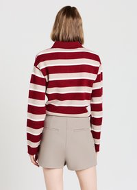 Gestreifter Pullover in Rot, Beige und Weiß; mit hohem Kragen; kombiniert mit hellbeigen Shorts mit Gesäßtaschen.