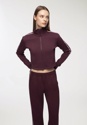 Tuta viola con giacca corta a zip e pantaloni abbinati. Presenta righe laterali bianche e una texture morbida con polsini e vita elastici.
