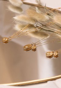 Goldene Halsketten mit verschiedenen Anhängern, die komplexe Gravuren und Texturen aufweisen und sich auf einer glatten Spiegeloberfläche spiegeln.