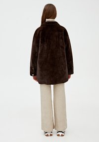 Veste en fausse fourrure marron avec un col, des manches longues et des accents de boutons, associée à un pantalon large beige et des baskets.