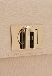 Dettaglio della borsa Furla con un esterno beige texturizzato, hardware color oro con un fermo logo e cuciture bianche lungo il bordo.