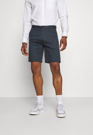 Navy shorts with a straight cut, button closure, and side pockets, paired with a white long-sleeve shirt and white socks with sneakers.