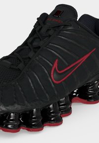 Μαύρο παπούτσι Nike Shox με κόκκινο λογότυπο swoosh και αναρτητικά ελατήρια στην σόλα, εμφανιζόμενο από κοντινή γωνιακή οπτική.