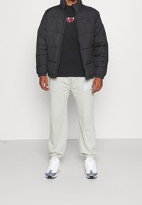 Giacca imbottita nera con collo alto, indossata sopra una t-shirt nera con stampa grafica. Pantaloni della tuta grigio chiaro con testo bianco. Scarpe da ginnastica bianche e grigie.