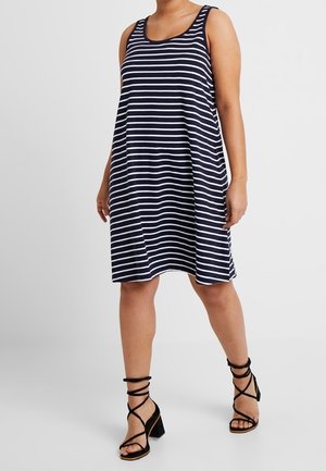 Vrouw draagt een mouwloze navyblauwe en witte jurk met horizontale strepen tot op de knie, met zwarte vetersandalen met blokhak.