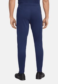 Marineblauwe sportbroek met een aansluitend ontwerp, elastische tailleband, zijritsen bij de zoom en een gladde textuur. Zwarte sneakers aan de voeten.