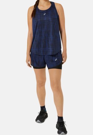 Vrouw die een donkerblauw patroon hardlooptanktopje en korte broek draagt, met zwarte sportschoenen en sokken, staand tegen een effen achtergrond.