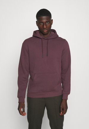 Giovane uomo con i capelli corti, indossa un hoodie bordeaux e pantaloni scuri, in piedi contro uno sfondo chiaro e semplice.