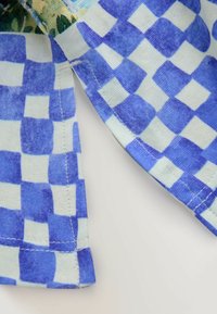 Blau-weißer karierter Stoff mit weicher Textur, der an der Kante eine Naht aufweist und ein teilweise sichtbares, bedrucktes Blumenmuster darüber hat.