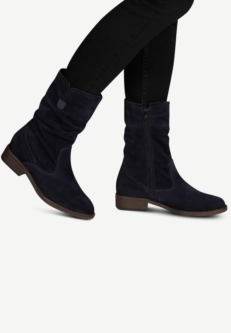 Bottes mid-calf en daim bleu marine foncé, souples, avec des talons bas en bois et des fermetures éclair latérales, portées avec un pantalon noir ajusté.