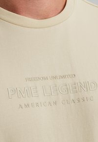 Camiseta de manga corta beige con gráficos en relieve: "LIBERTAD ILIMITADA", "LEYENDA PME" y "CLÁSICO AMERICANO". Fabricada en suave algodón.