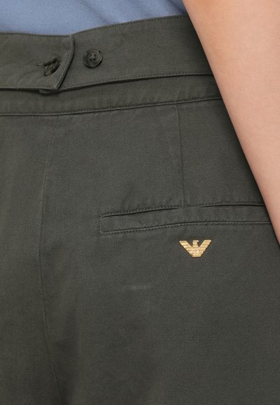 Σκούρο πράσινο παντελόνι από υφασμάτινη υφή, με μπροστινή τσέπη και χρυσό λογότυπο κοντά στη μέση. Δύο μαύρα κουμπιά ορατά.