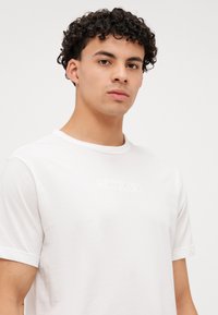 Bijela pamučna majica kratkih rukava s diskretnim reljefnim logotipom Calvin Klein sprijeda. Klasični izrez oko vrata.