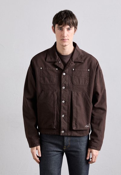 Veste en coton marron avec un col, deux poches poitrine et des boutons en métal argenté. Son design présente une coupe décontractée et un ourlet droit, associée à un jean noir.