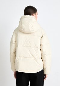 Puffer jacket in light cream with a detachable hood, featuring quilted, padded sections and a relaxed fit, worn over black pants.