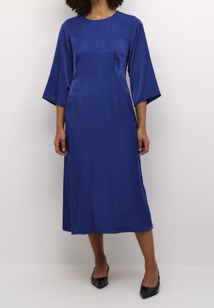 Femme portant une robe bleu cobalt mi-longue avec des manches larges trois-quarts et des escarpins noirs à bout pointu, debout devant un fond uni.