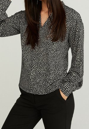 Kvinde iført en sort og hvid polkaprikket bluse med lange ærmer og sorte bukser, den ene hånd i lommen, håret løst over skuldrene.