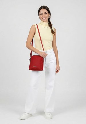Woman with braided hair wearing a sleeveless yellow turtleneck, white pants, white sneakers, and carrying a red crossbody bag.