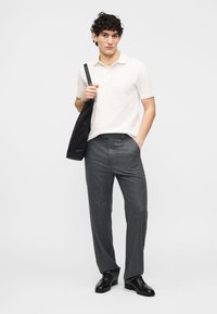 Νεαρός άνδρας που φοράει λευκό κοντομάνικο πόλο, γκρι παντελόνια, μαύρα παπούτσια, κρατώντας μαύρη τσάντα ώμου, στέκεται μπροστά σε απλό ανοιχτό φόντο.