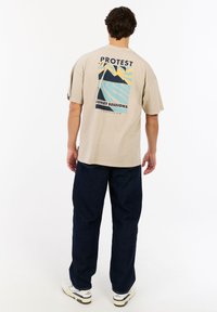 Beige t-shirt met korte mouwen met een kleurrijke grafische print op de achterkant, featuring ontwerpen van bergen en golven. Gecombineerd met donkerblauwe broek.