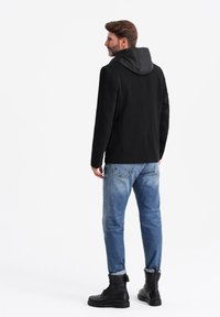 Svart kappa med vadderad huva, skräddarsydd form och slät textur, i kombination med blå jeans och svarta kängor.