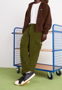 Person med brun zip-up fleecejacka, vit t-shirt, olivgröna corduroy cargobyxor och svarta sneakers lutar sig mot en blå metallförvaringsvagn.