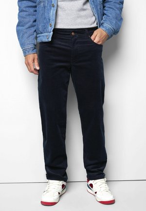 Mann trägt schwarze Hose, weiße Sneakers mit roten und blauen Akzenten, graues Shirt und blaue Jeansjacke mit einer Hand in der Tasche.
