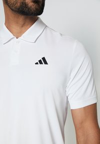 Polo blanc en tissu lisse et léger, avec un logo Adidas noir sur la poitrine gauche. Boutons au col.