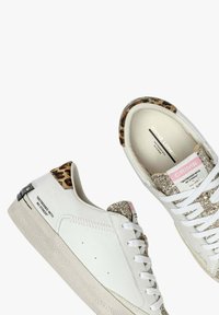 Zapatillas de piel blanca con suela texturizada, con un talón de estampado de leopardo, acentos brillantes y el texto "Desgastadas sin estrés".