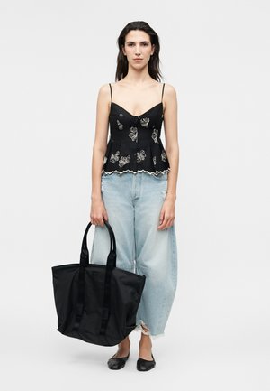 Femeie purtând un top negru floral cu bretele spaghetti și blugi deschiși la culoare cu croială largă, ținând o geantă mare tote neagră și încălțată cu pantofi negri comozi.