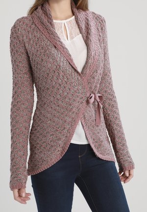 Roze gebreide omslagtrui met een gestructureerd patroon en striksluiting, gedragen over een wit topje met kanten rand en gecombineerd met donkerblauwe jeans.