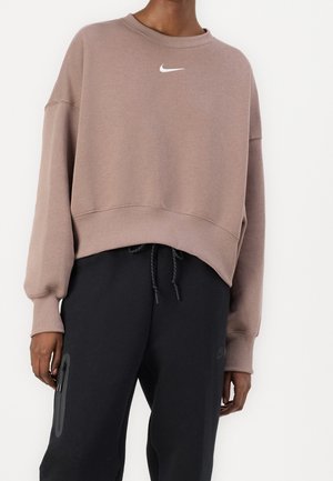 Osoba nosząca przyciętą taupe bluzę Nike z białym logo i czarne spodnie ze sznurkiem oraz detalem kieszeni na zamek.