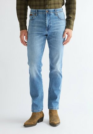 Man wearing light blue jeans, brown suede ankle boots, and a green plaid long-sleeve shirt, standing against a plain background.