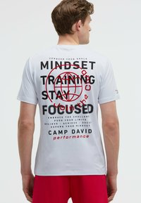 White short-sleeve T-shirt featuring bold black and red text on the back, with motivational phrases and a circular graphic.