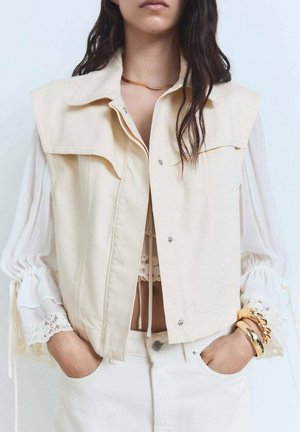 Femme portant un gilet beige sans manches sur une blouse blanche avec des poignets en dentelle, un pantalon blanc, des bracelets en or et un collier en or, les mains dans les poches.
