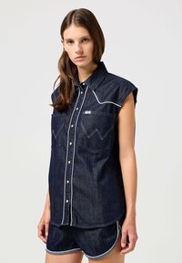 Denim skjorte med korte ermer, mørk vask, hvit piping langs kragen og lommene, med to brystlommer og knapper foran.