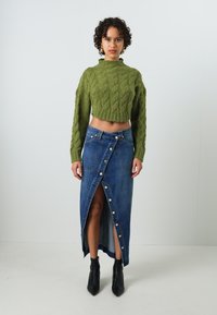 Pull en maille torsadée vert court associé à une jupe en denim bleu avec une fermeture à boutons à l'avant et une fente haute. Bottines à talons noires.