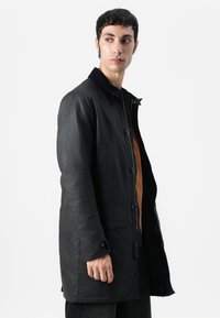 Manteau noir d'extérieur avec une finition texturée, dotée d'une fermeture à boutons, de deux poches latérales et d'un col contrastant.
