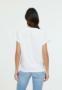 T-shirt en coton blanc avec des manches courtes roulées, col rond et ourlet droit. L'arrière-plan est uni, mettant en avant la simplicité du t-shirt.