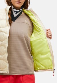 Veste matelassée beige avec un intérieur vert vif, portée sur un haut texturé marron. Une écharpe noire à pois blancs est également visible.