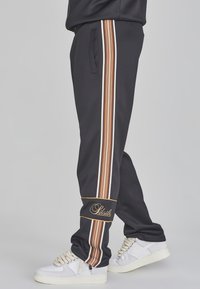 Zwarte sportbroek met verticale strepen in oranje, bruin en wit, zijzakken en een logo aan de onderkant. Gecombineerd met witte sneakers.