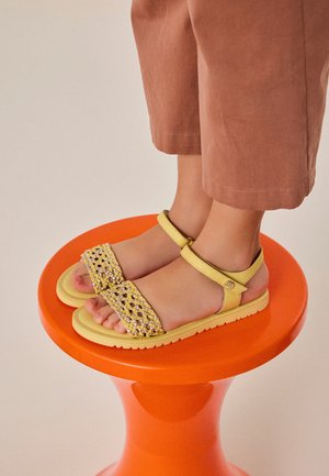 Niño con sandalias amarillas de tiras tejidas de pie sobre un taburete redondo de color naranja brillante contra un fondo neutro.