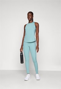 Lichtblauw sporttanktop en leggings van ademend materiaal, gecombineerd met witte sneakers en met een zwarte waterfles in de hand.